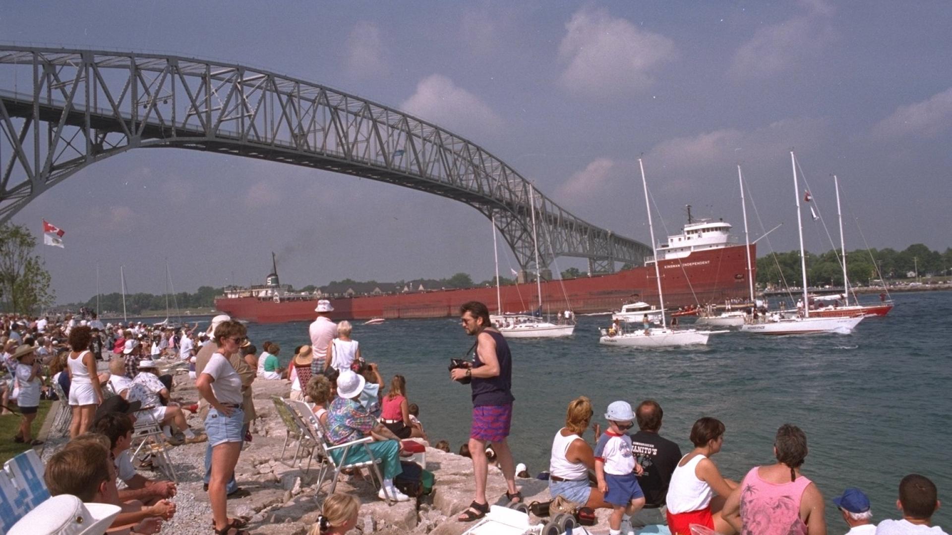 People sitting by the shore as a cargo ship goes under the bridge.