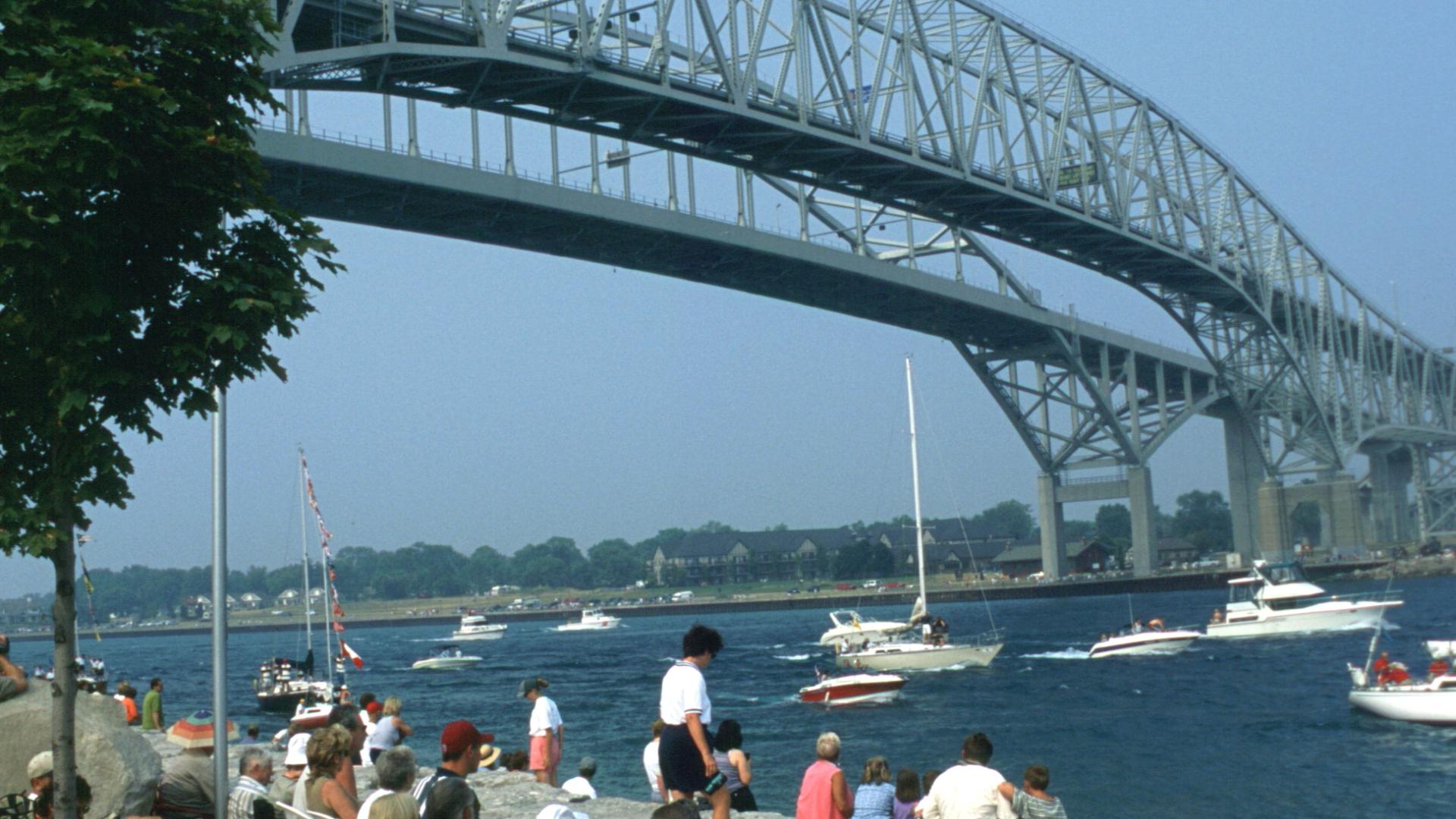 People sitting at the bay watching as boats sail under the bridge.