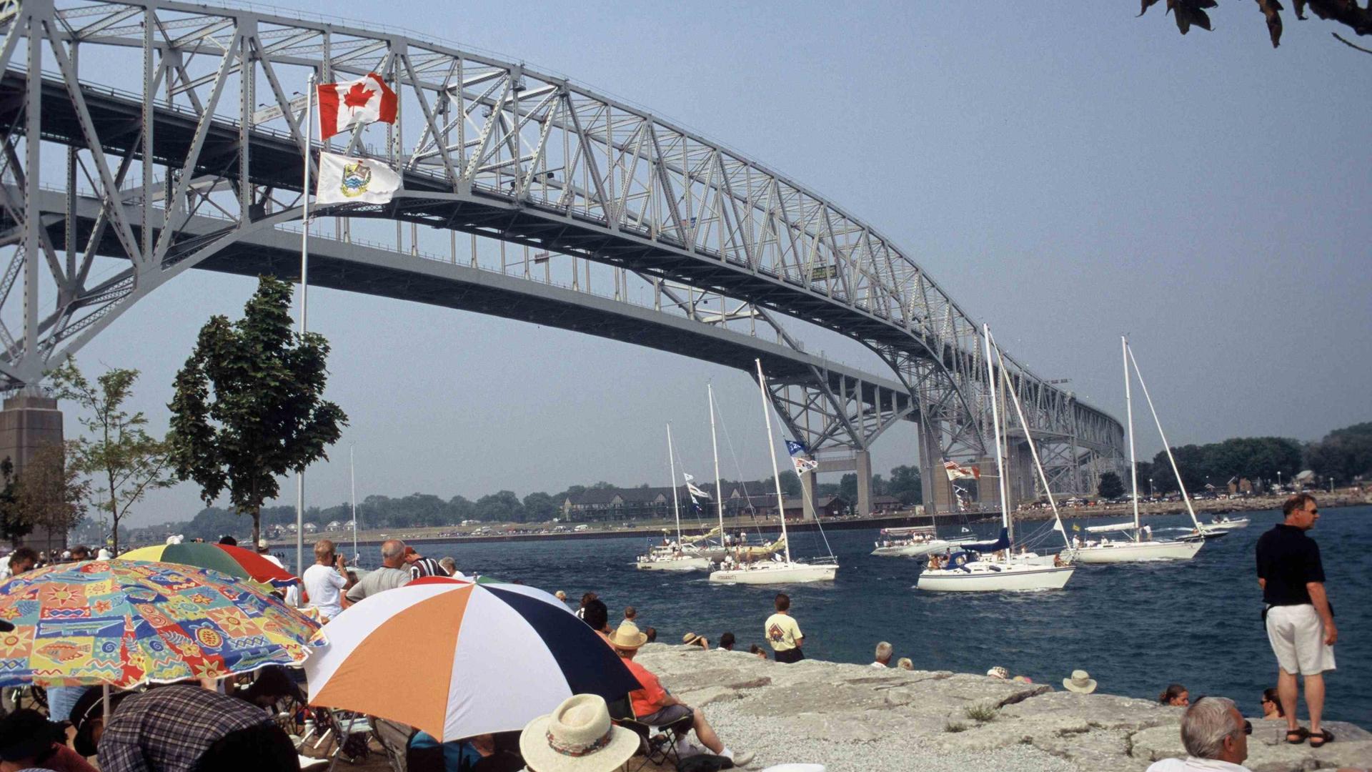 People sitting by the shore watching as boats sail by under bridge.