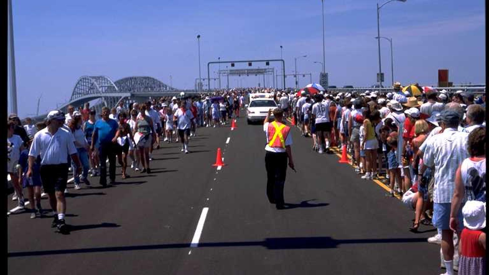 Massive group of people standing on sides of the bridge. A person is controlling traffic with a flag.