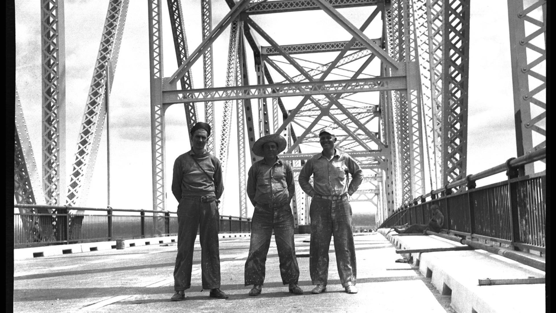 Three men standing on completed section of bridge. 