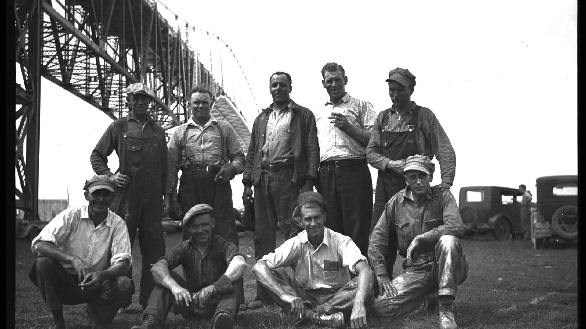 Group of men posing in front of constructed bridge. The bridge stands tall on the background.