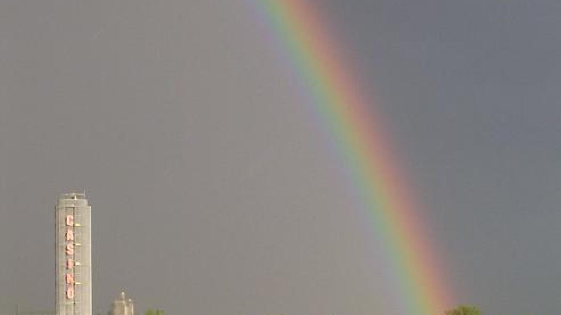 Rainbow sprouting from the ground. There are some buildings on the foreground. The rainbow is well-defined/visible.