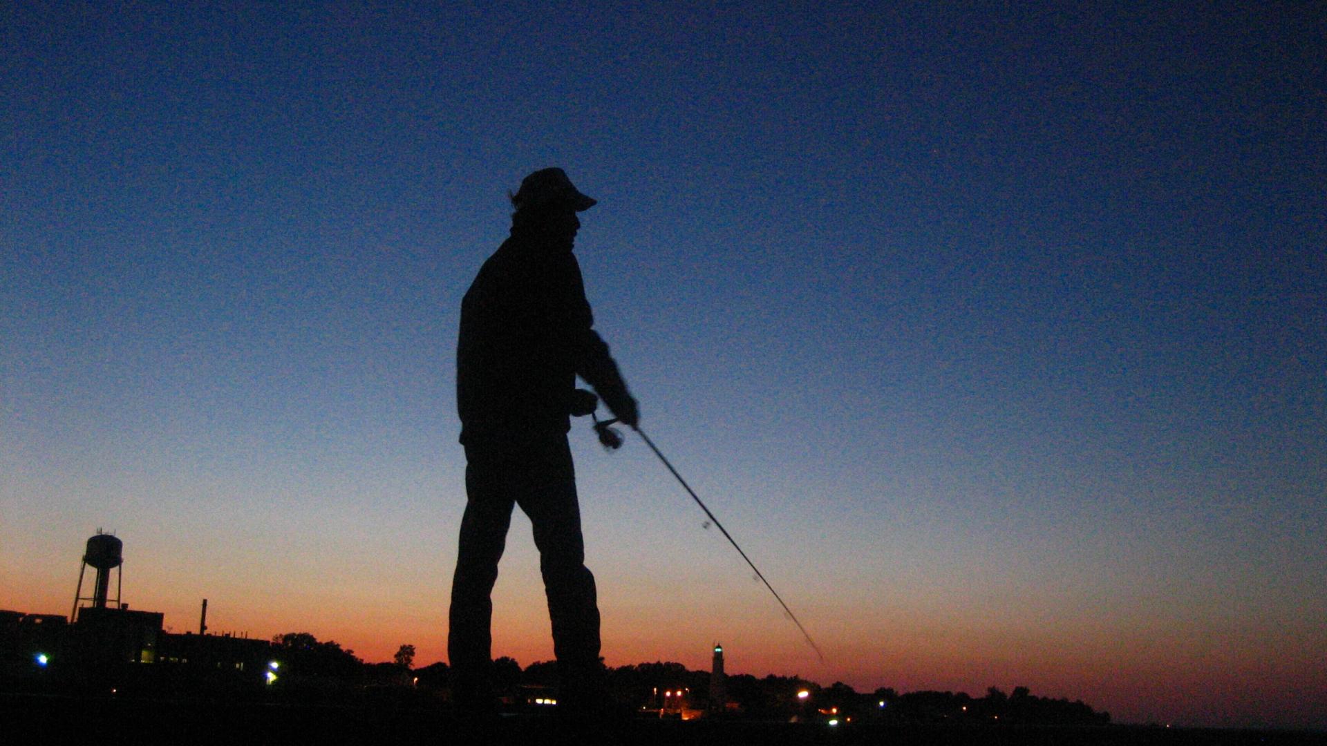 Silhouette of a fisherman fishing. The background is a gradient of dark blue on the top and orange near the horizon. 