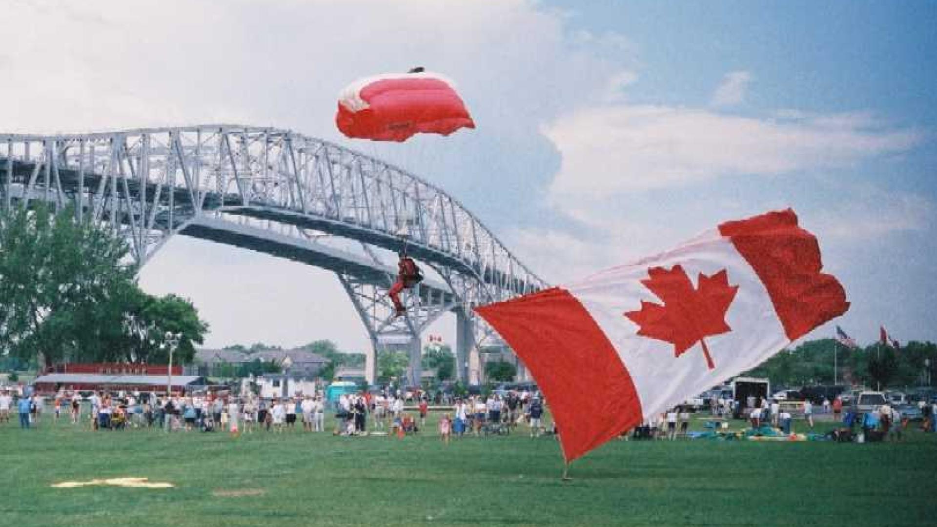 Parachuter landing on green patch of grass. The parachuter has a Canadian flag parachute design. The bridge stands tall in the background.