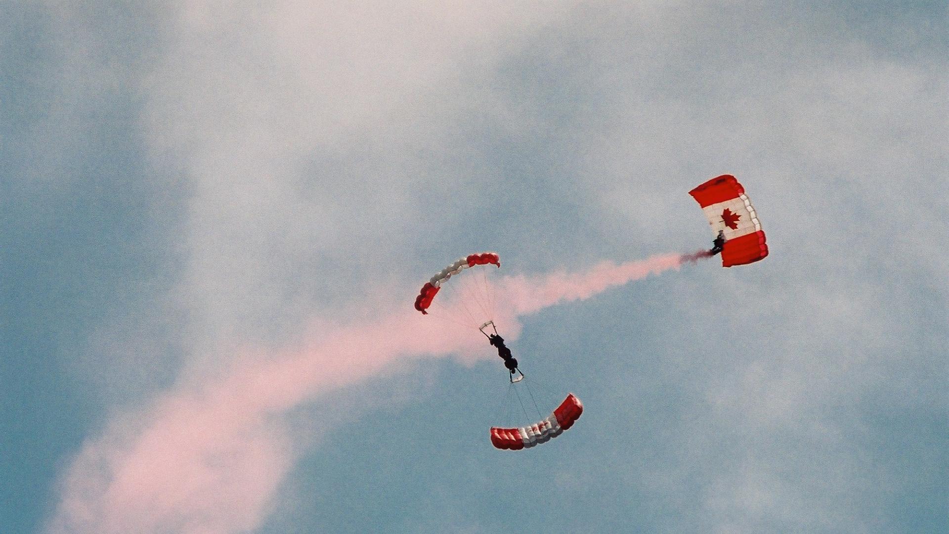 Parachuter sailing through the sky. The parachute has a Canadian flag design. The parachuter is leaving a red trail of smoke behind.