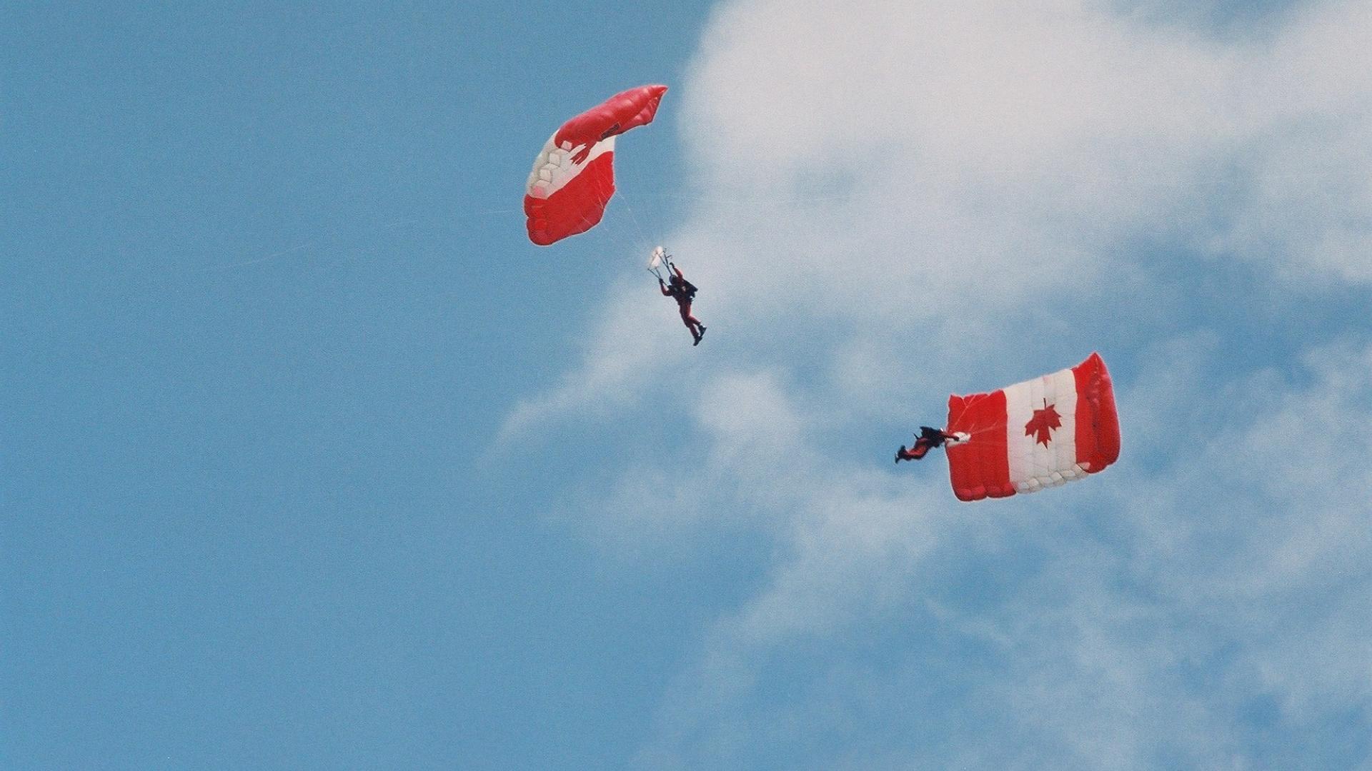 Parachuter sailing through the sky. The parachute has a Canadian flag design. The parachuter is waving the Canadian flag.