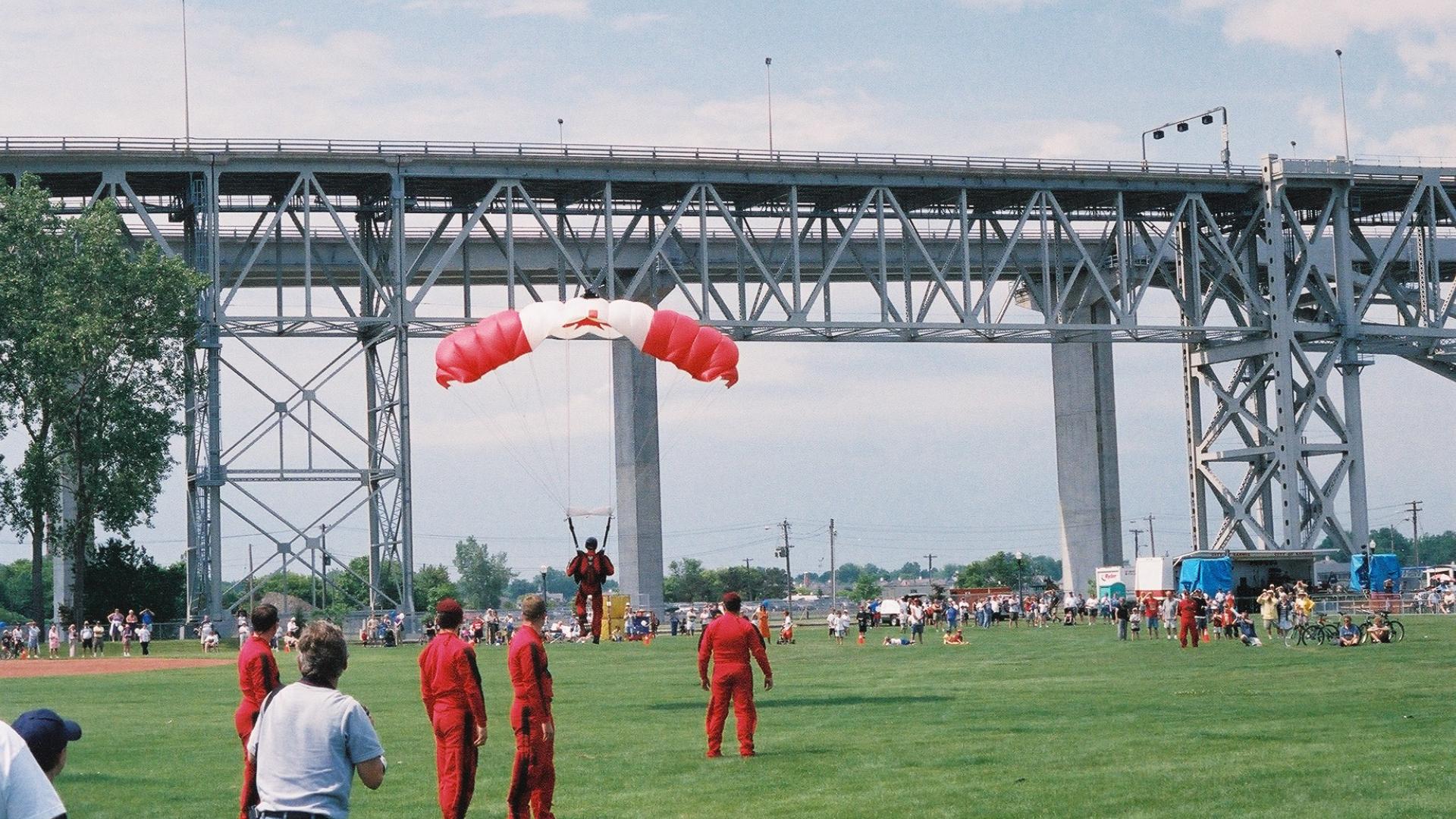 Parachuter landing on green patch of grass. The parachuter has a Canadian flag parachute design. The bridge stands tall in the background.