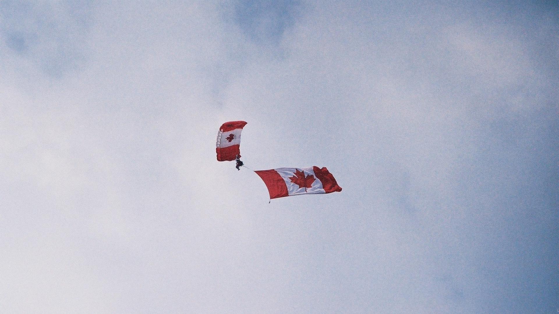 Person parachuting. The parachute has the flag of Canada design. The parachuter is waving the Canadian flag.