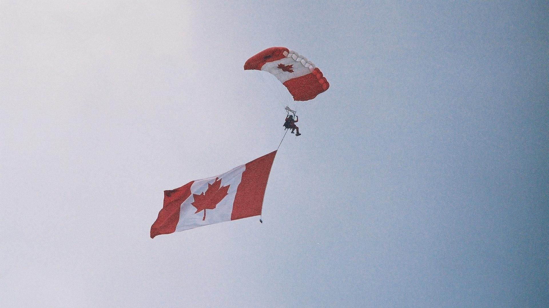 Person parachuting. The parachute has the flag of Canada design. The parachuter is waving the Canadian flag.
