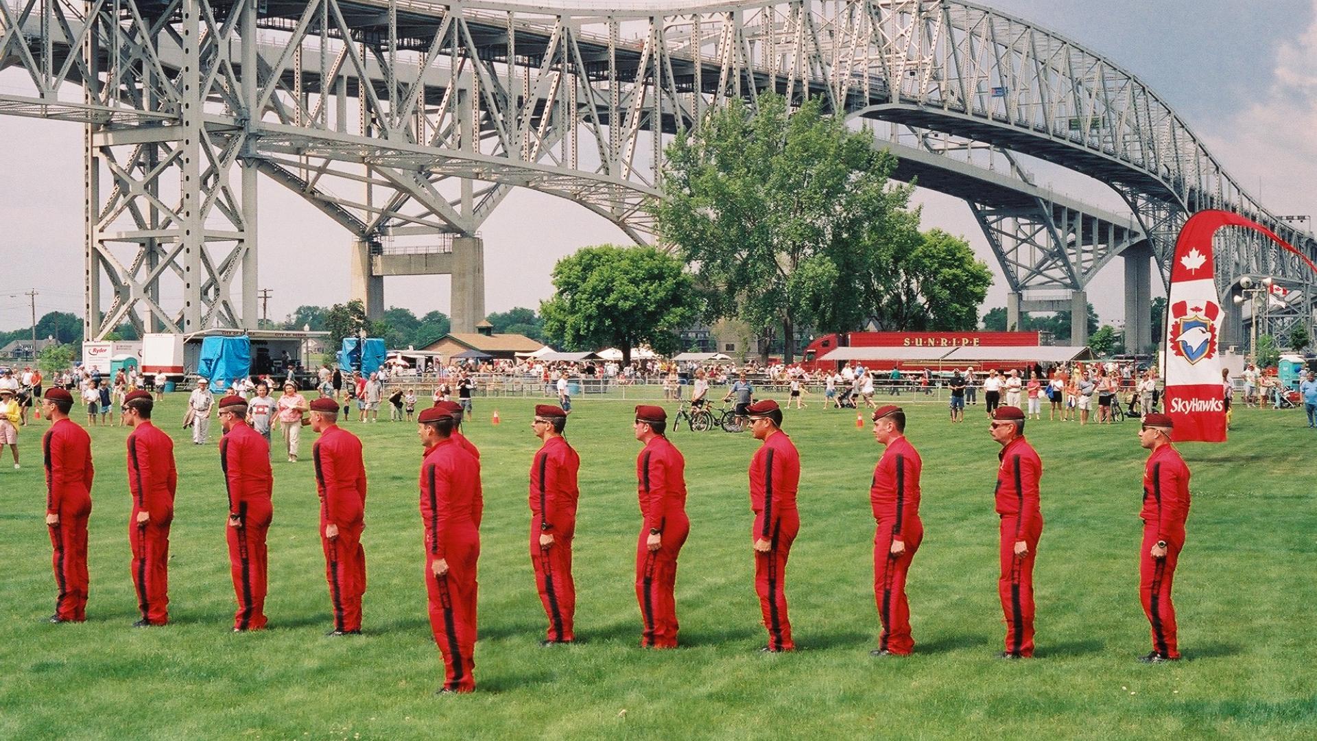Group of men dressed in red jumpsuits standing in attention. The bridge stands tall in the background.