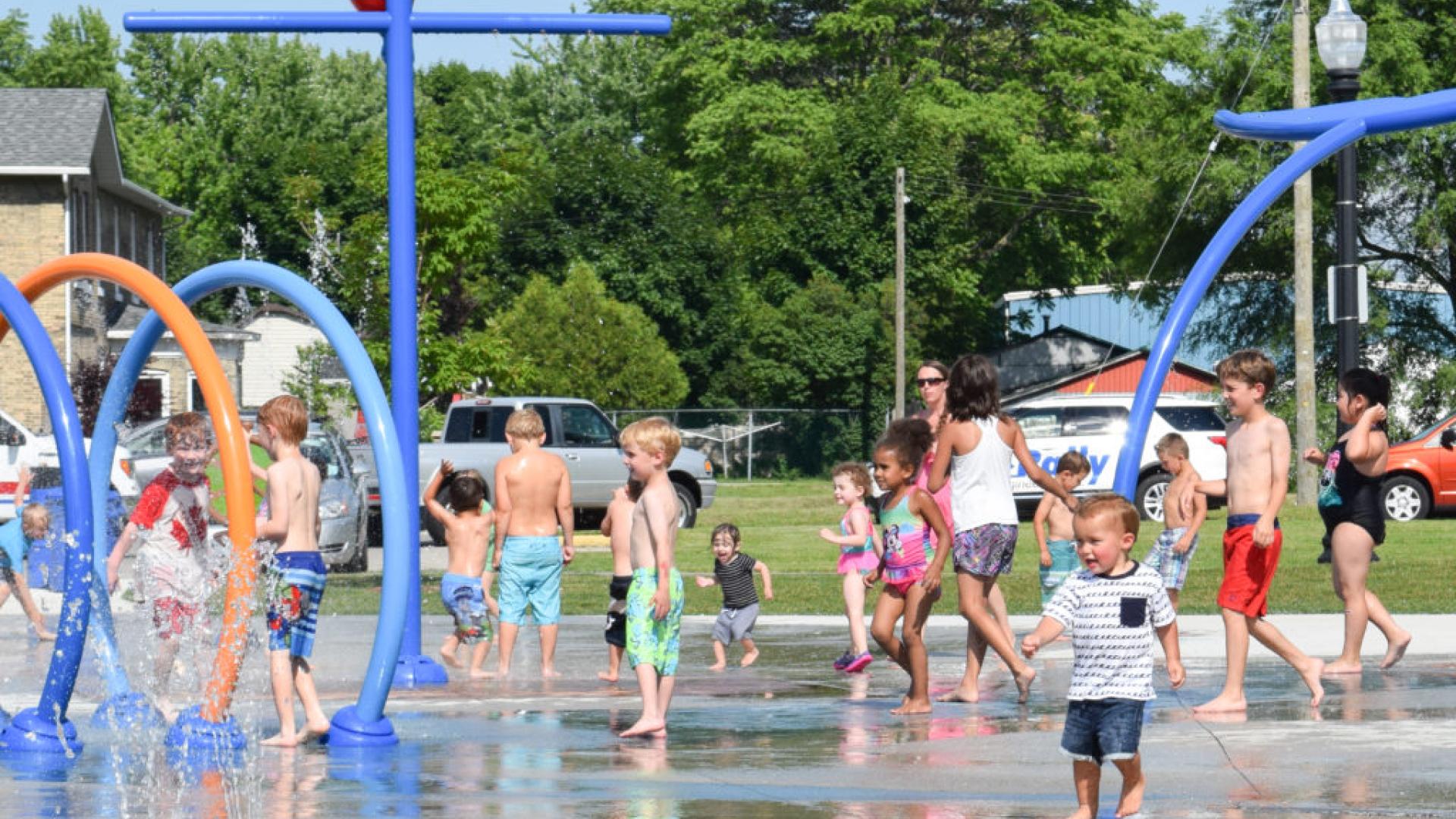Children playing in a splashpark. There are a random assortment of water pipes and spouts.