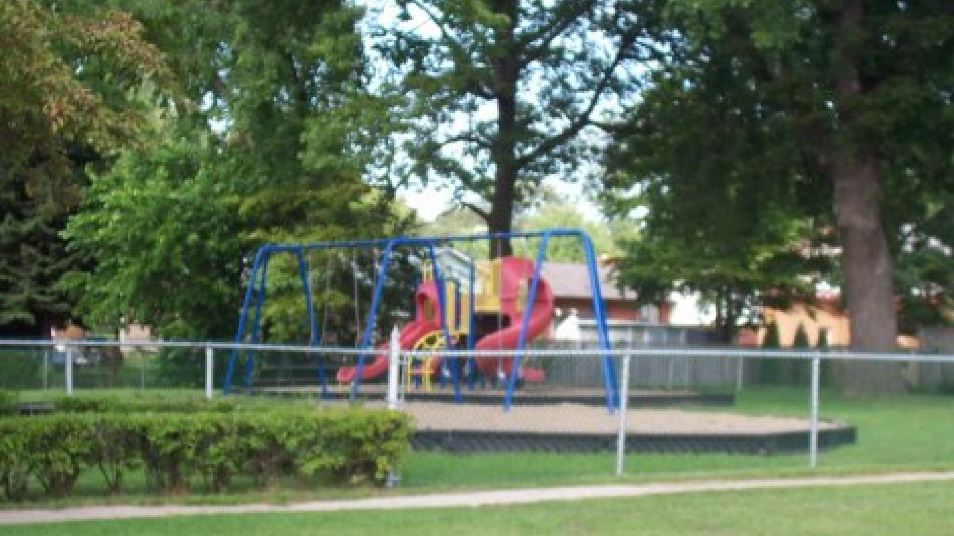 Playground with  a junglge gym and a swing set. There is a fence in the foreground and some trees in the background..