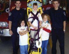 Child strapped to upright gurney with neck brace smiling flanked by 2 other children with neckbraces. there are also two fire marshals flanking the child in the gurney.