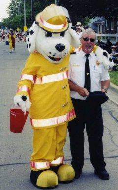 Fire chief standing next to a Dalmatian mascot dressed in a yellow firesuit.