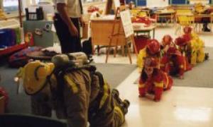 Row of children in classroom mimicking a firefighter crawling on the floor as a fire cheif observes them.